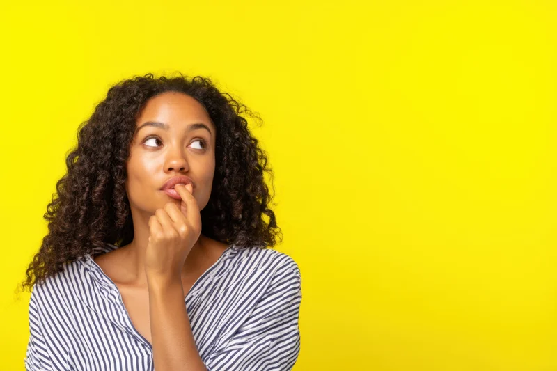 Young woman with curly hair biting her lip thoughtfully against yellow background
