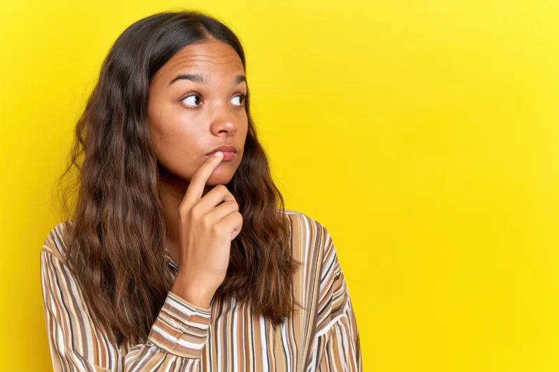 Young woman with thoughtful expression biting her lip against yellow background