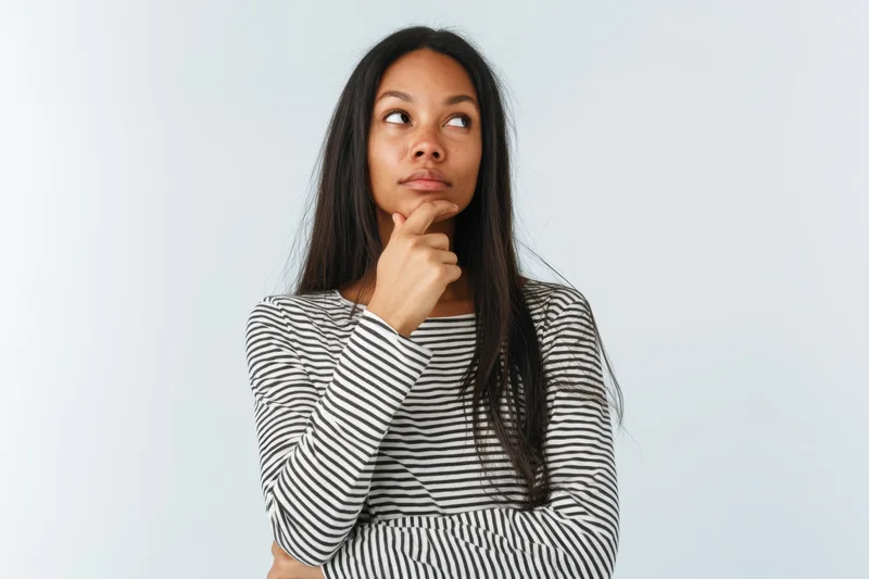 Young woman with long dark hair wearing striped longsleeve shirt, hand on chin, looking upward in thought against white background