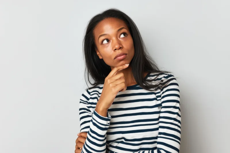 Young black woman in striped longsleeve shirt thinking with hand on chin against white background
