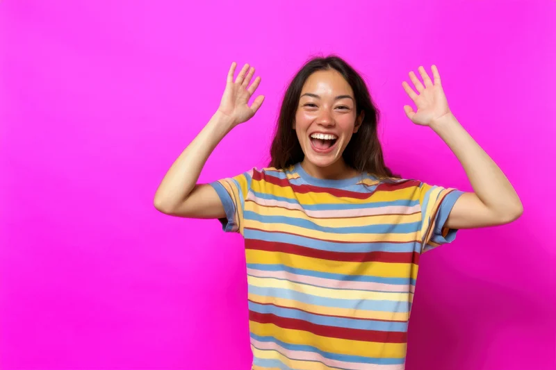 Young asian woman laughing with arms raised in front of pink background wearing colorful striped shirt