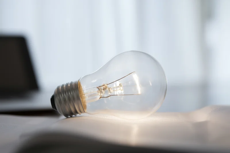 Illuminated lightbulb resting on open book with laptop in background