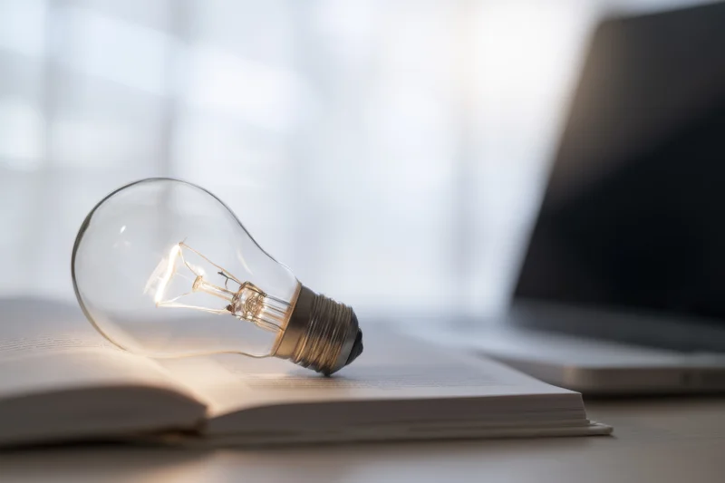 Illuminated lightbulb resting on open book on desk near laptop