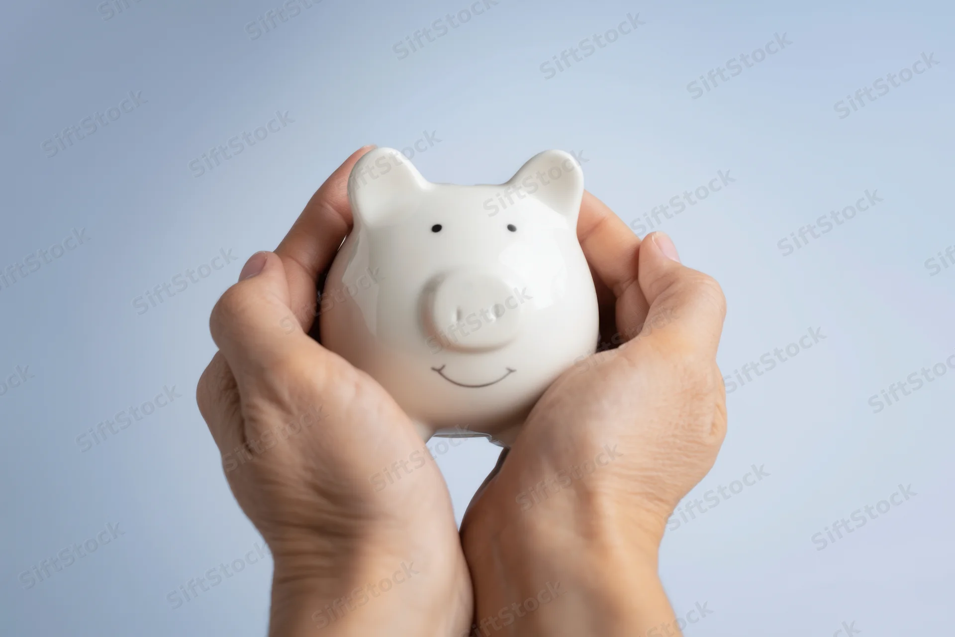 Hands gently holding a white ceramic piggy bank with a cheerful face against a plain background