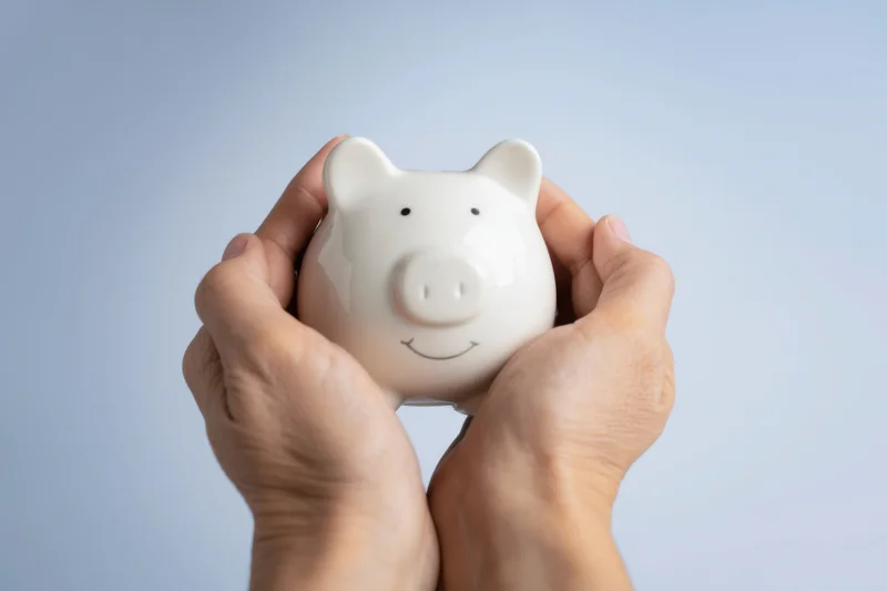 Hands gently holding a white ceramic piggy bank with a cheerful face against a plain background