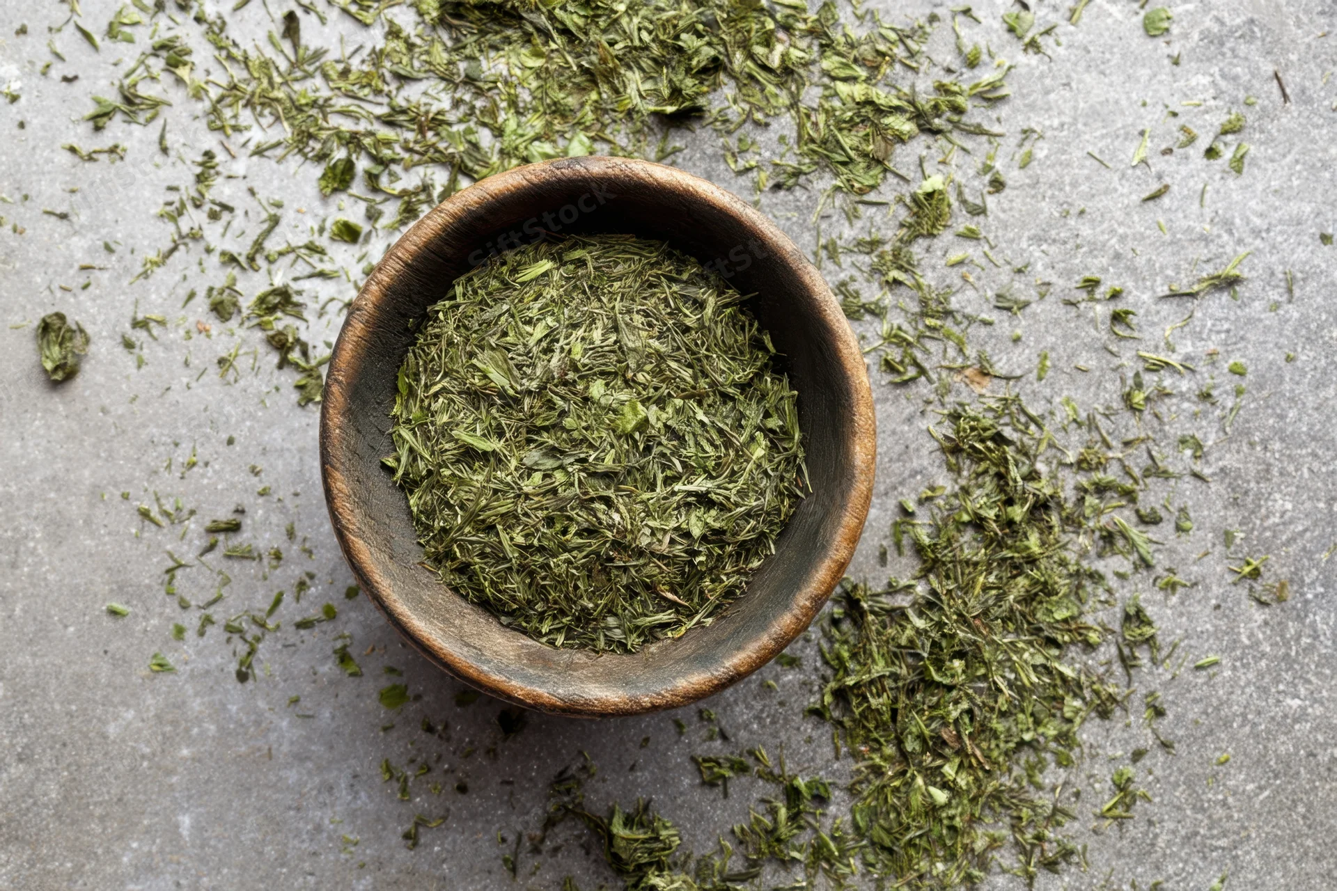 Loose green tea leaves in a rustic wooden bowl scattered on a gray stone surface
