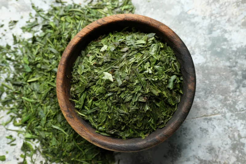 Loose green tea leaves in a wooden bowl scattered on a stone surface