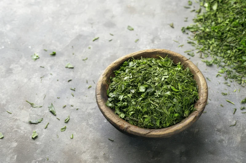 Loose green tea leaves in a wooden bowl scattered on a gray surface