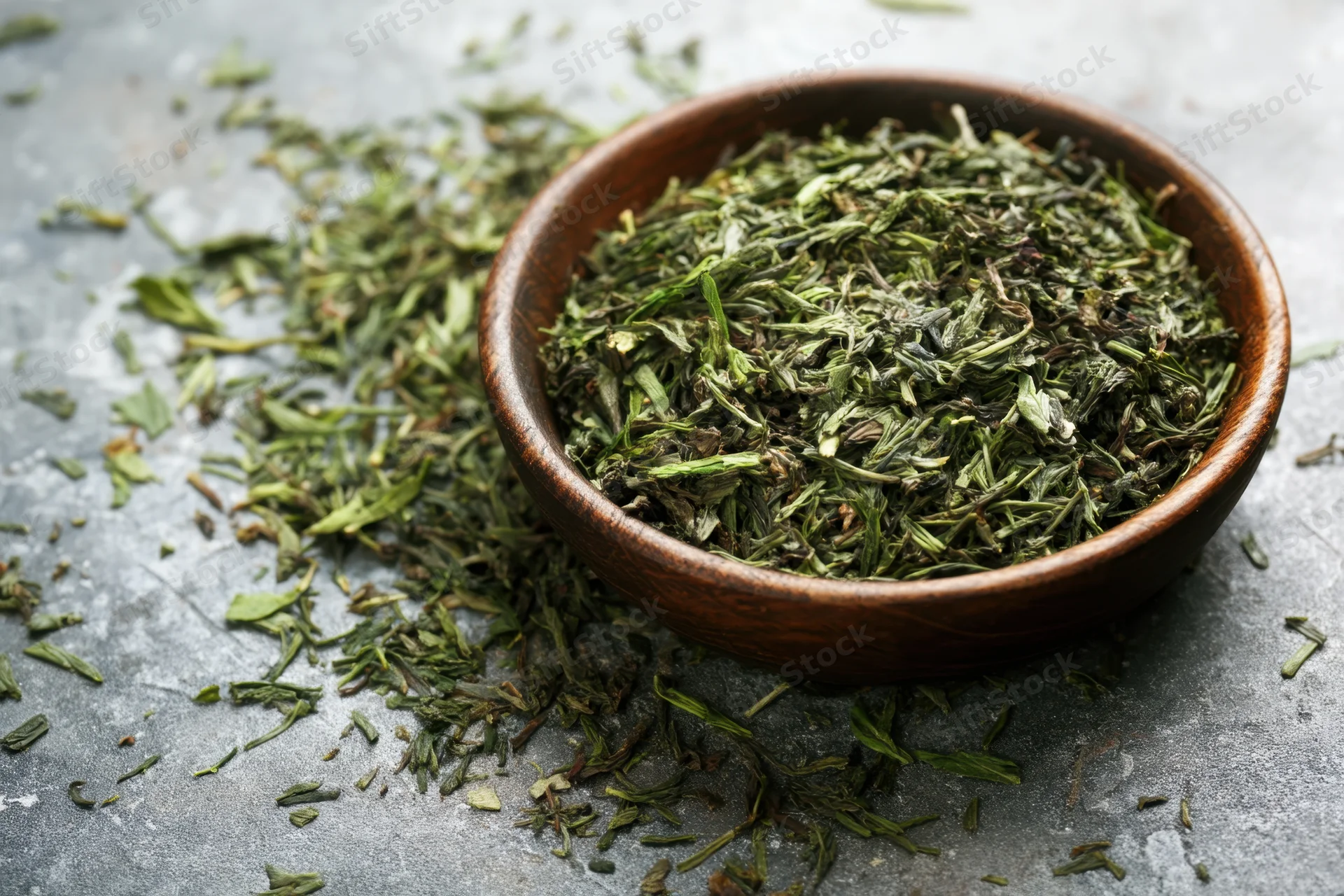 Loose green tea leaves in a wooden bowl scattered on a gray textured surface