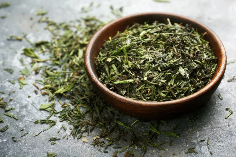 Loose green tea leaves in a wooden bowl scattered on a gray textured surface
