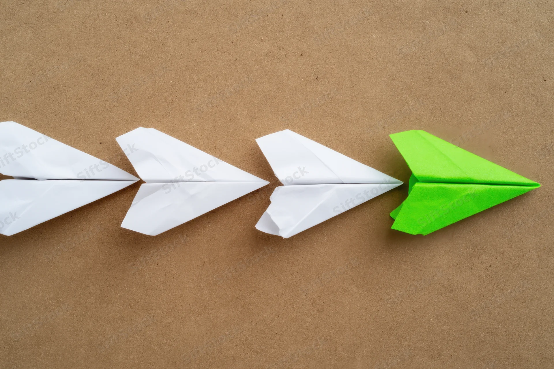 Four paper airplanes arranged in a line on a brown surface, three white and one green at the end