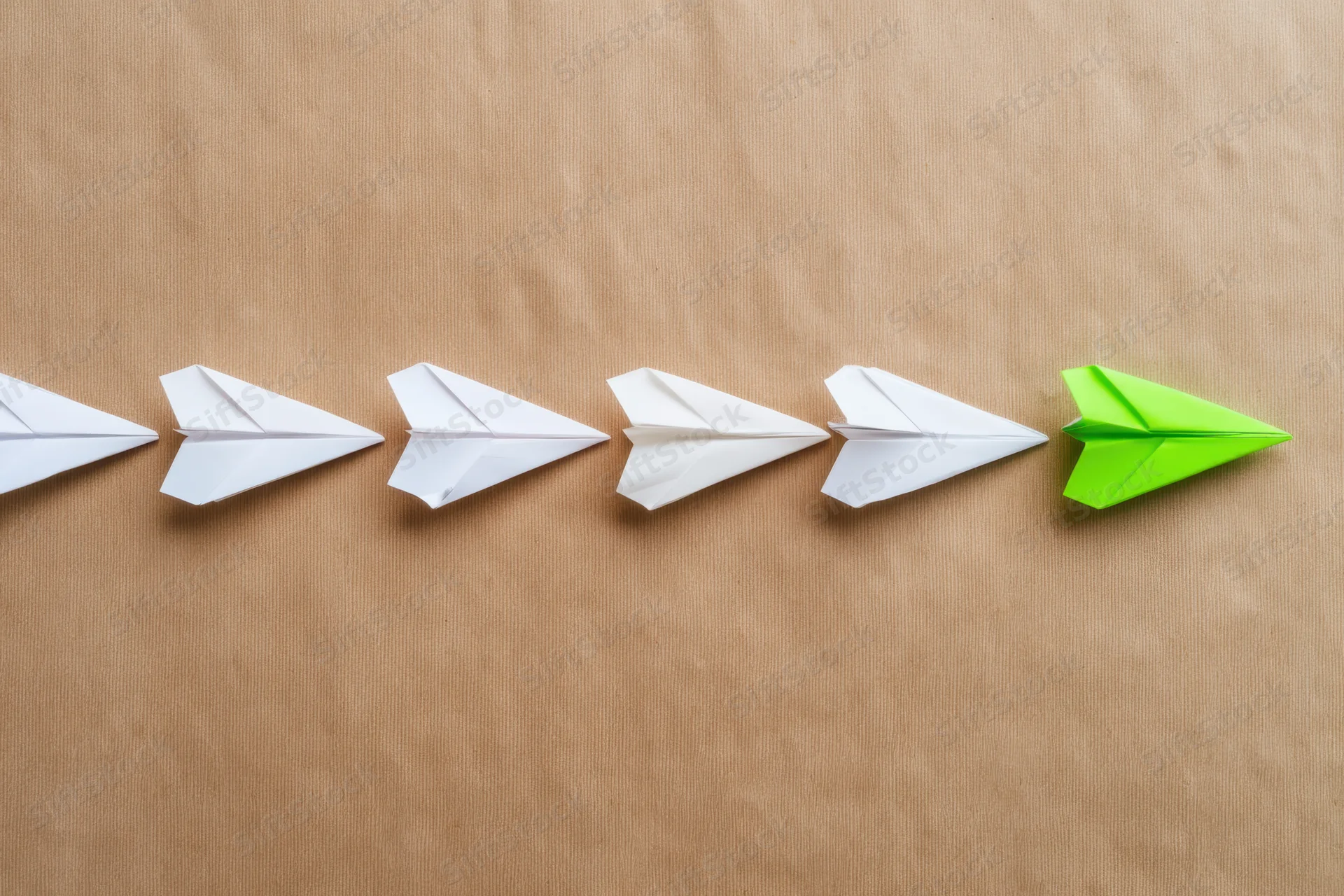 A row of white paper airplanes with one green airplane at the end on a brown surface