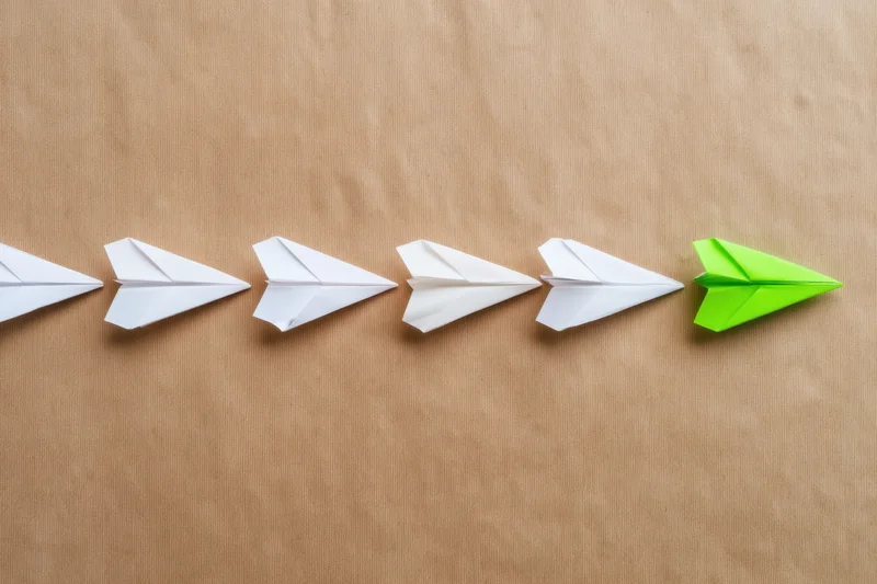 A row of white paper airplanes with one green airplane at the end on a brown surface