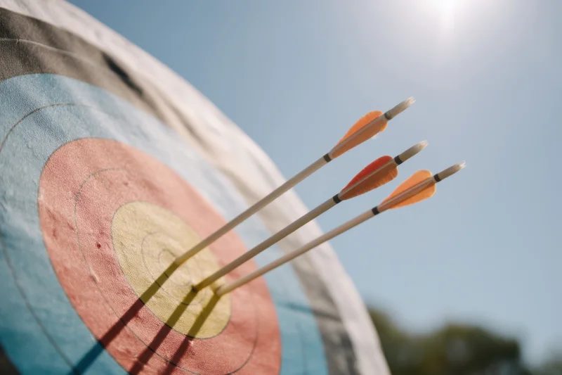 Three arrows embedded in the center of a colorful archery target under bright sunlight