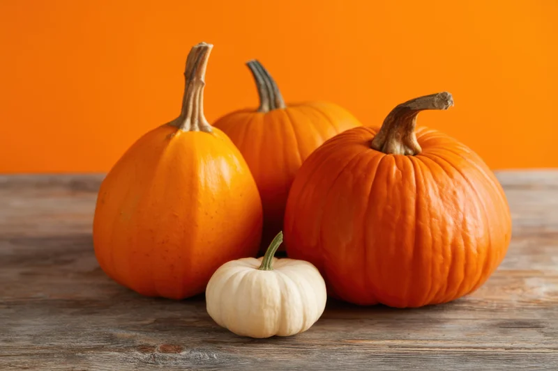 Three pumpkins of varying sizes and one small white pumpkin arranged on a wooden table against an orange background