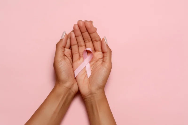 Two hands gently holding a pink awareness ribbon against a solid pink background