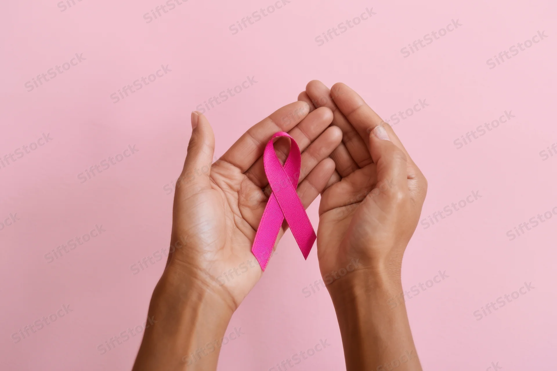 Hands gently holding a pink ribbon against a pink background