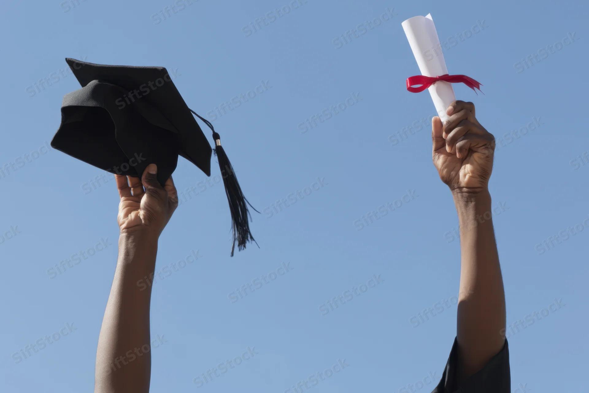 Hands holding graduation cap and diploma raised against clear blue sky