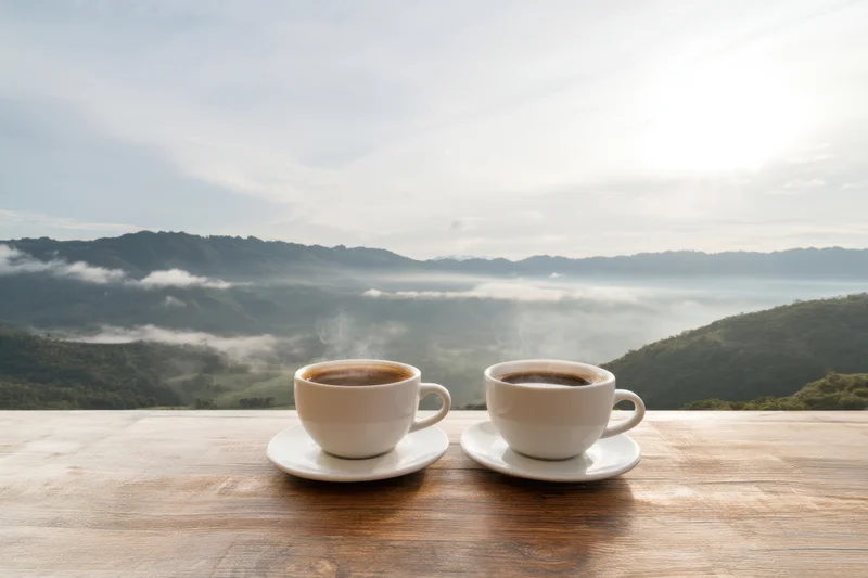 Two steaming white coffee cups on a wooden table overlooking a misty mountain valley at sunrise
