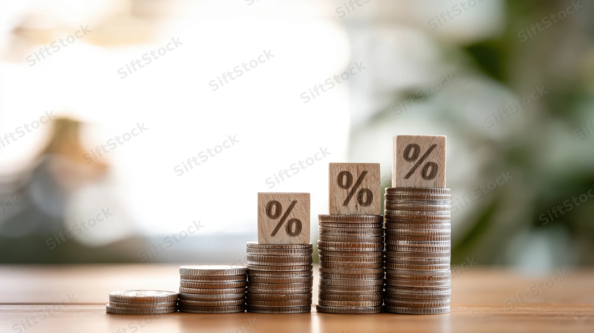 Four stacks of coins increasing in height each topped with a wooden block marked with a percentage symbol on a wooden table with blurred background