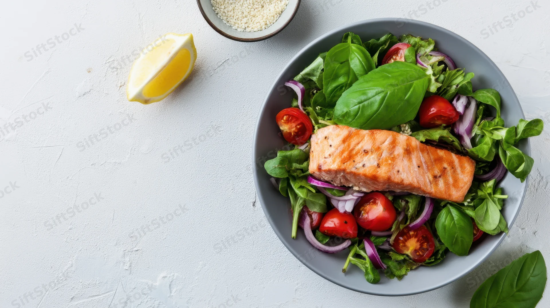 Salmon fillet atop a salad of greens, tomatoes, onions, and basil in a gray bowl on a white surface