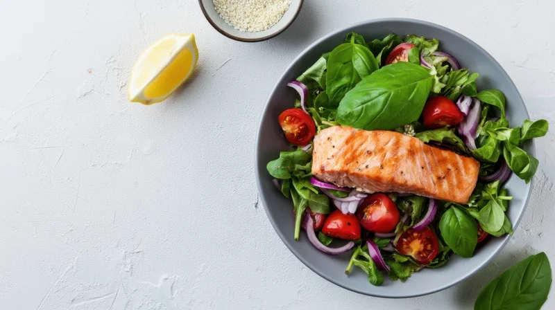 Salmon fillet atop a salad of greens, tomatoes, onions, and basil in a gray bowl on a white surface