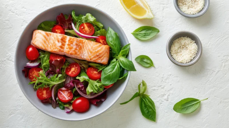 Salmon fillet atop a mixed green salad with cherry tomatoes, red onion, basil, and lemon wedge in a gray bowl on a white surface