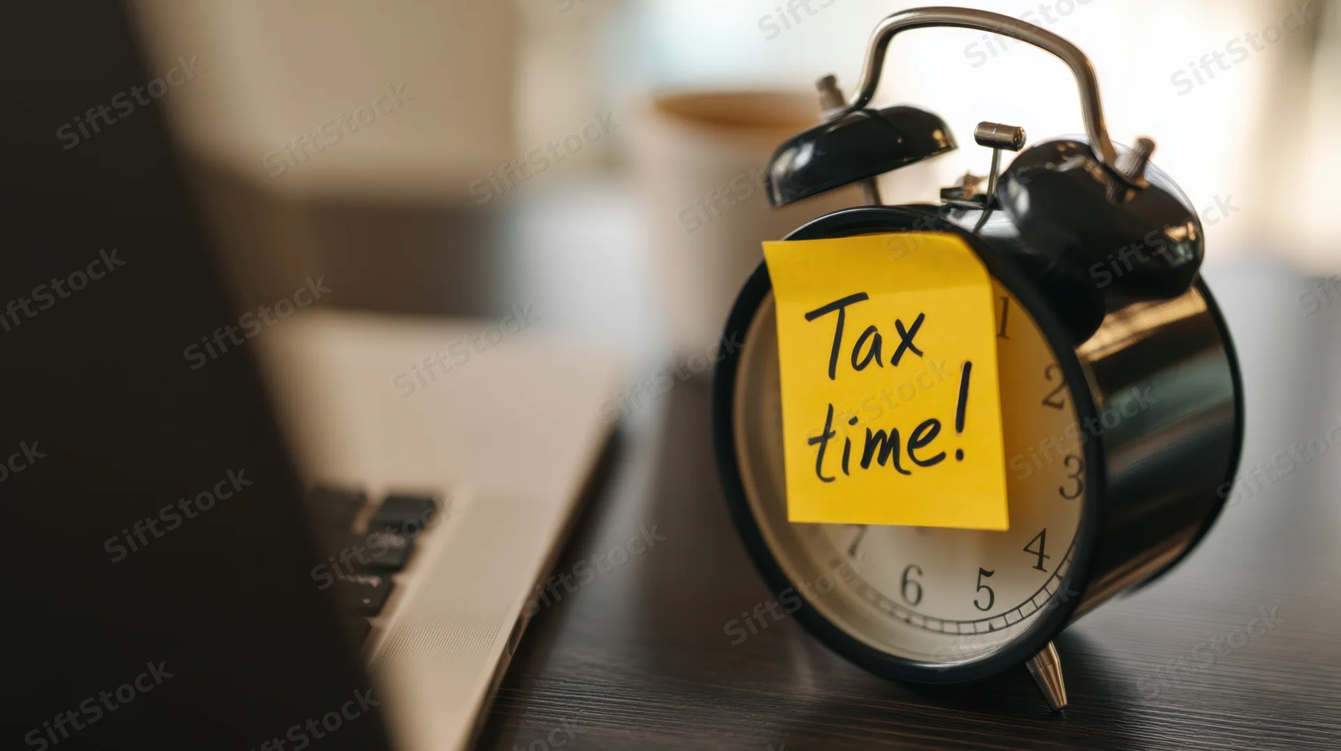 Classic alarm clock with yellow sticky note reading tax time on wooden desk beside laptop keyboard