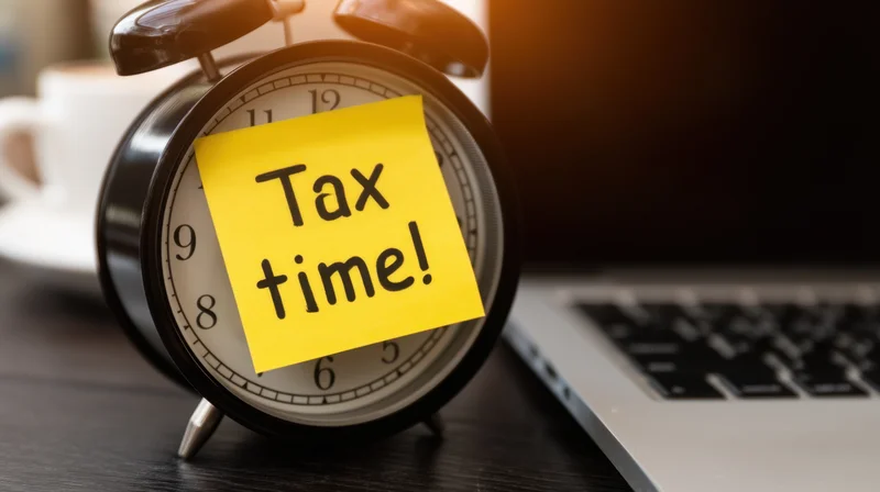 Classic alarm clock with yellow sticky note reading tax time on office desk beside laptop and coffee cup