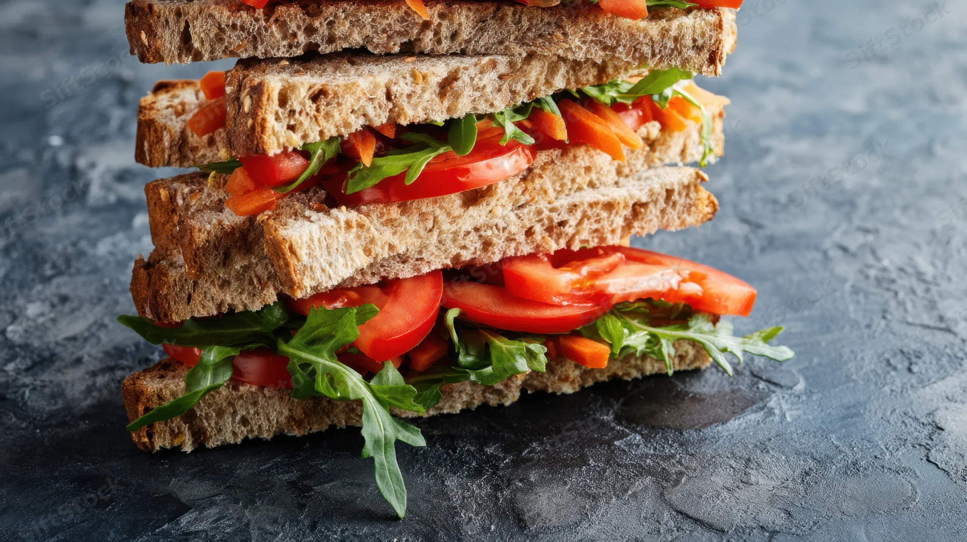 Stacked wholegrain sandwich with tomato arugula and carrot on dark stone surface