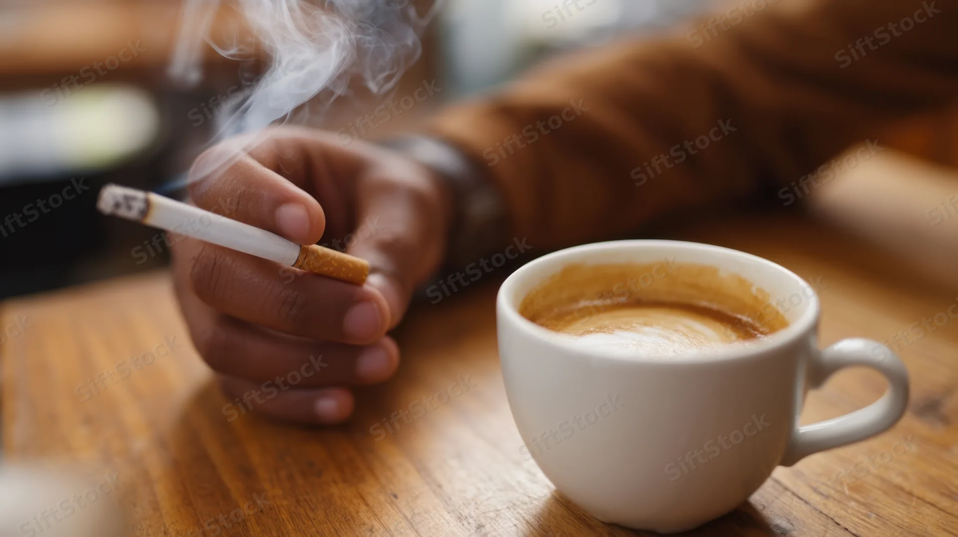 Hand holding a burning cigarette with smoke rising beside a white coffee cup on a wooden table