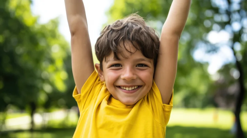 Young boy smiling with arms raised in a park wearing a yellow tshirt