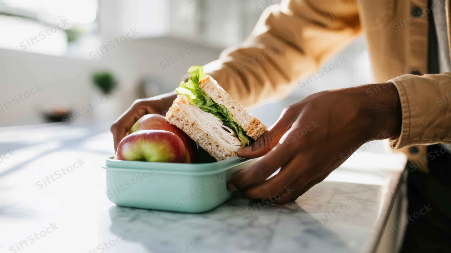 Hands placing a sandwich and two apples into a lunchbox on a kitchen countertop