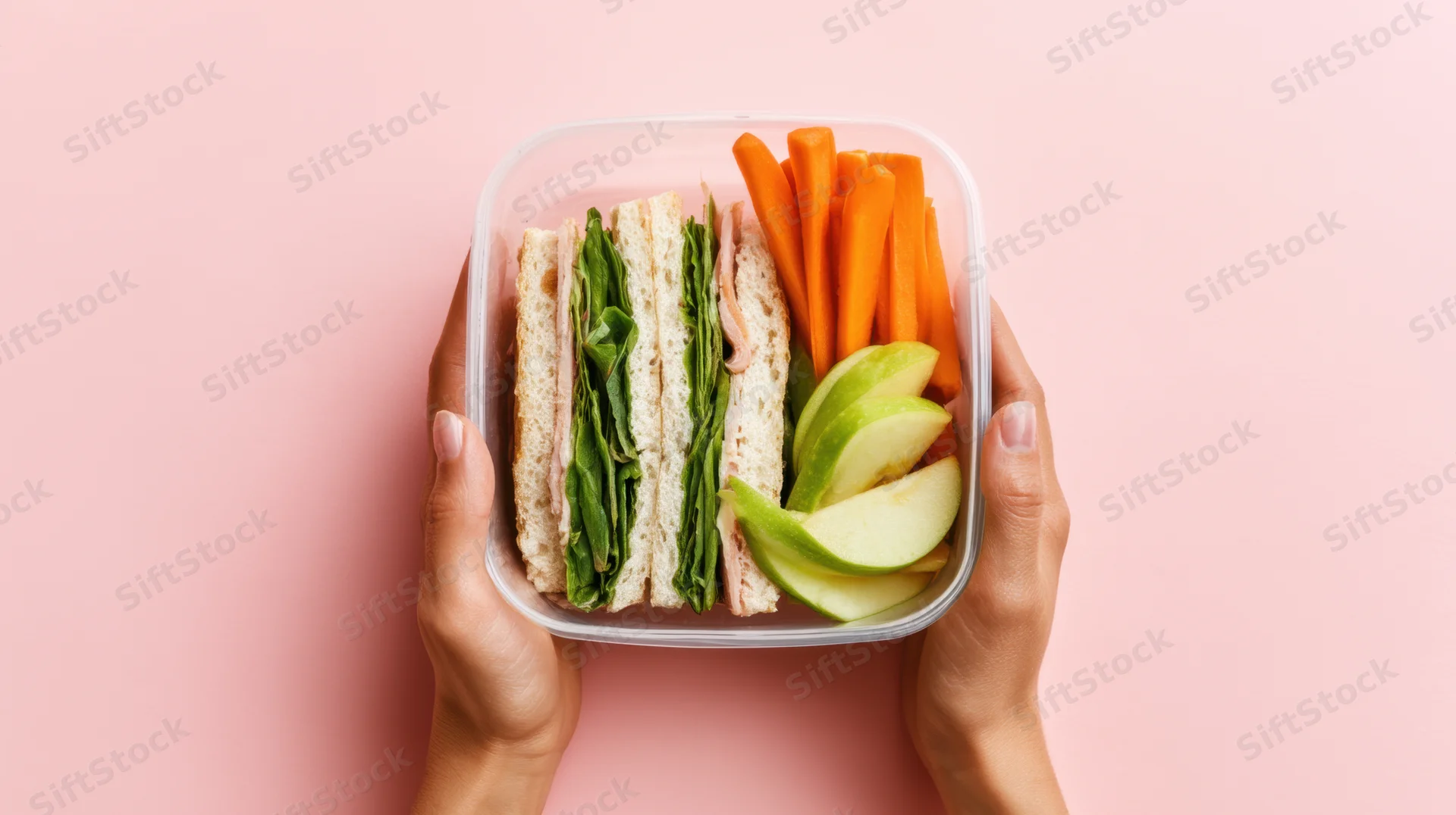 Hands holding a transparent lunchbox with a sandwich, carrot sticks, and apple slices against a pink background