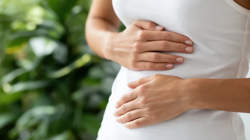Woman with hands on stomach in white tank top near green foliage
