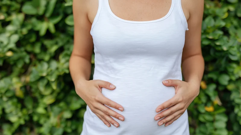 Woman in white tank top with hands resting on stomach outdoors near green foliage