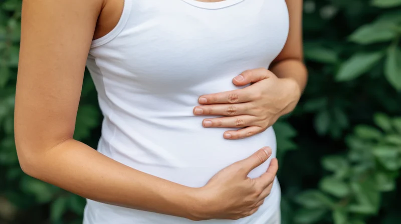 Woman in white tank top with hands gently resting on her stomach outdoors among green foliage
