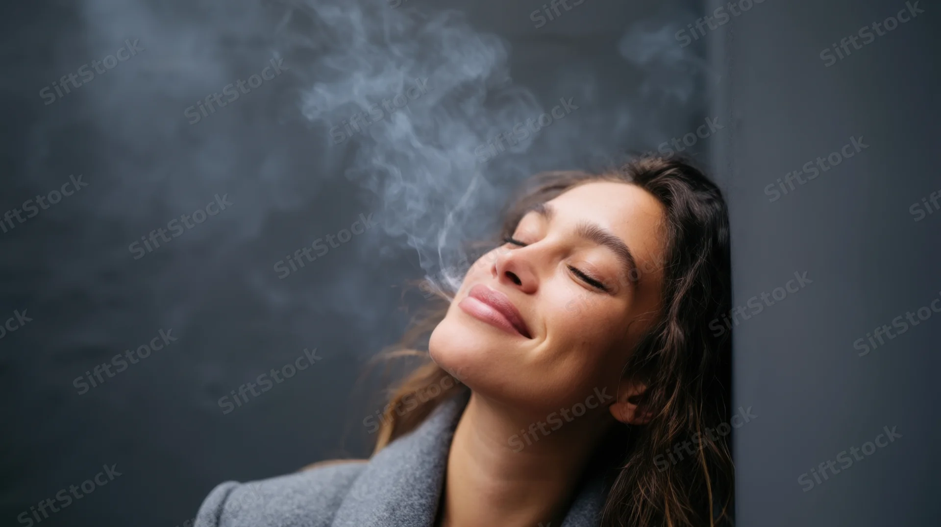 Woman leaning against dark gray wall exhaling smoke with eyes closed