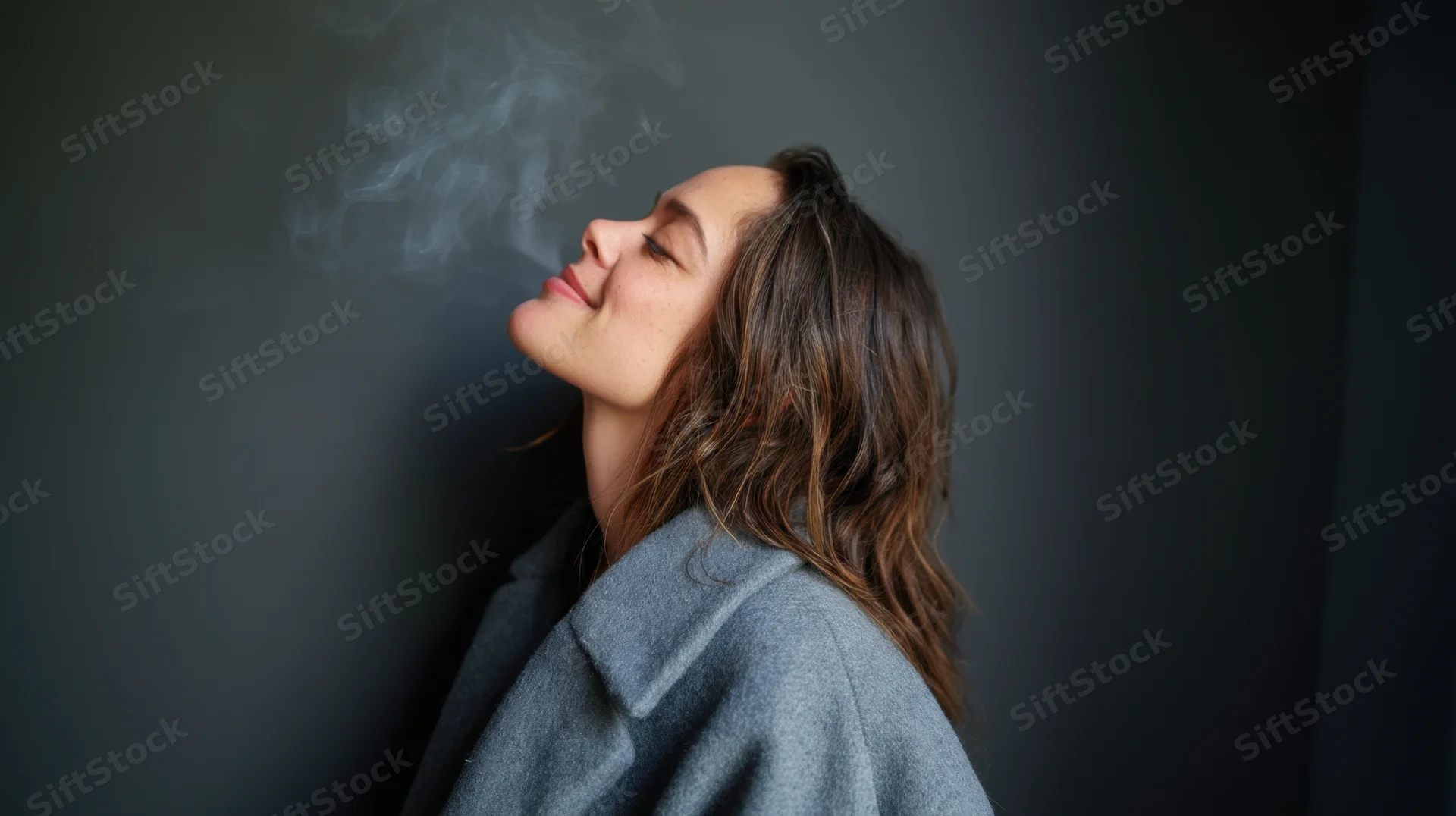 Woman leaning against dark gray wall exhaling smoke with eyes closed in profile view