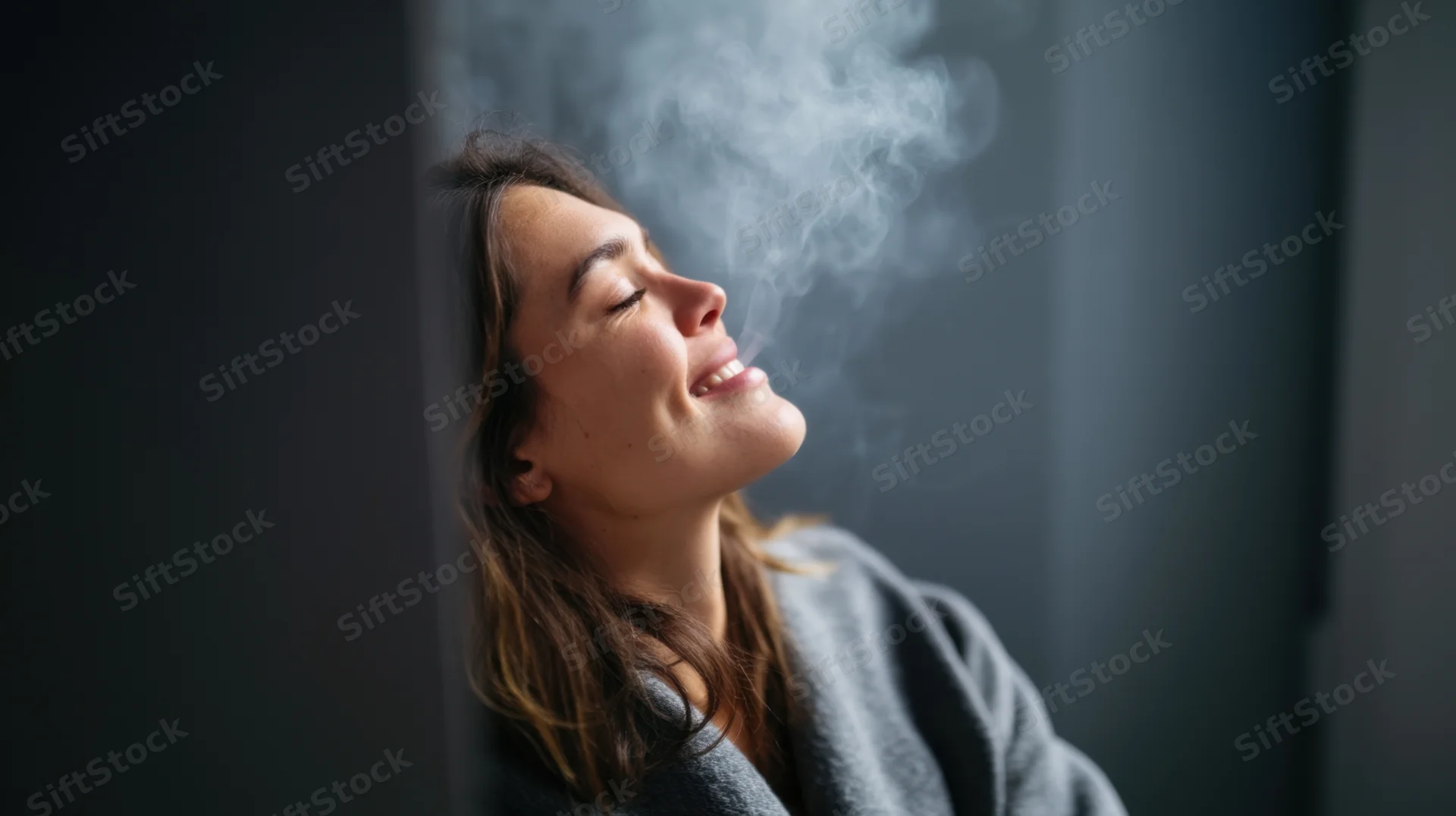 Woman exhaling smoke while leaning against a dark gray wall, smiling with eyes closed, wearing a robe
