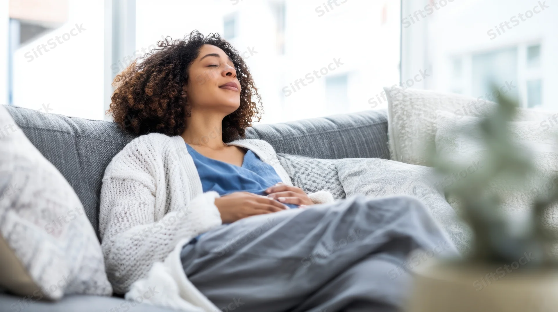 Woman resting with eyes closed on a gray couch in a bright living room