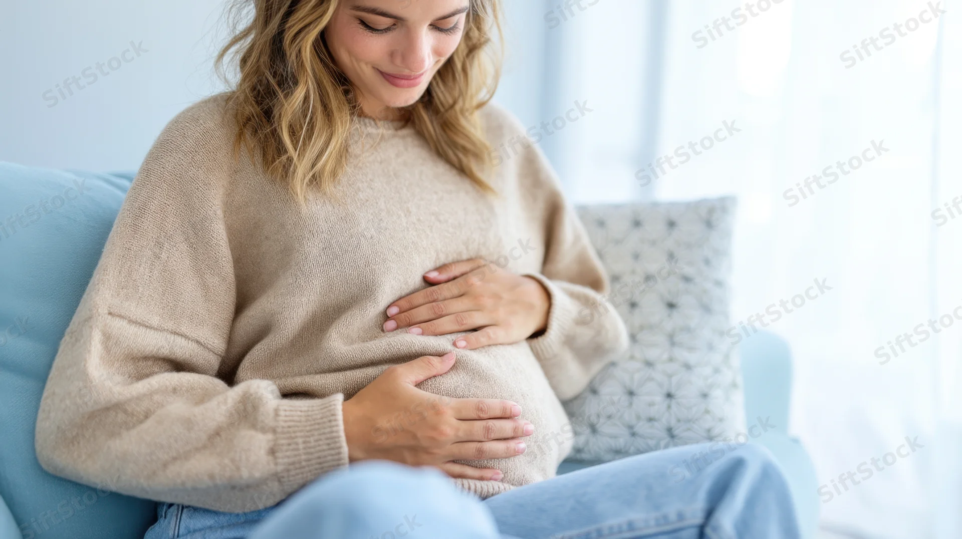 Pregnant woman sitting on a blue couch with hands on her belly, smiling softly in a bright living room