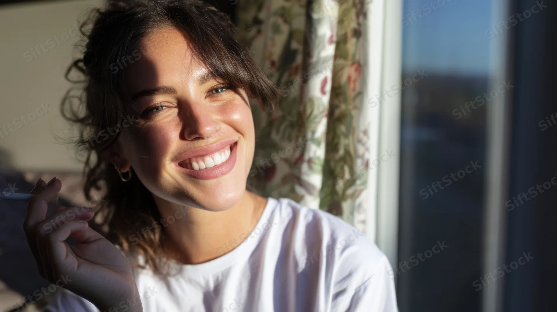 Woman smiling while holding a cigarette near a sunlit window indoors