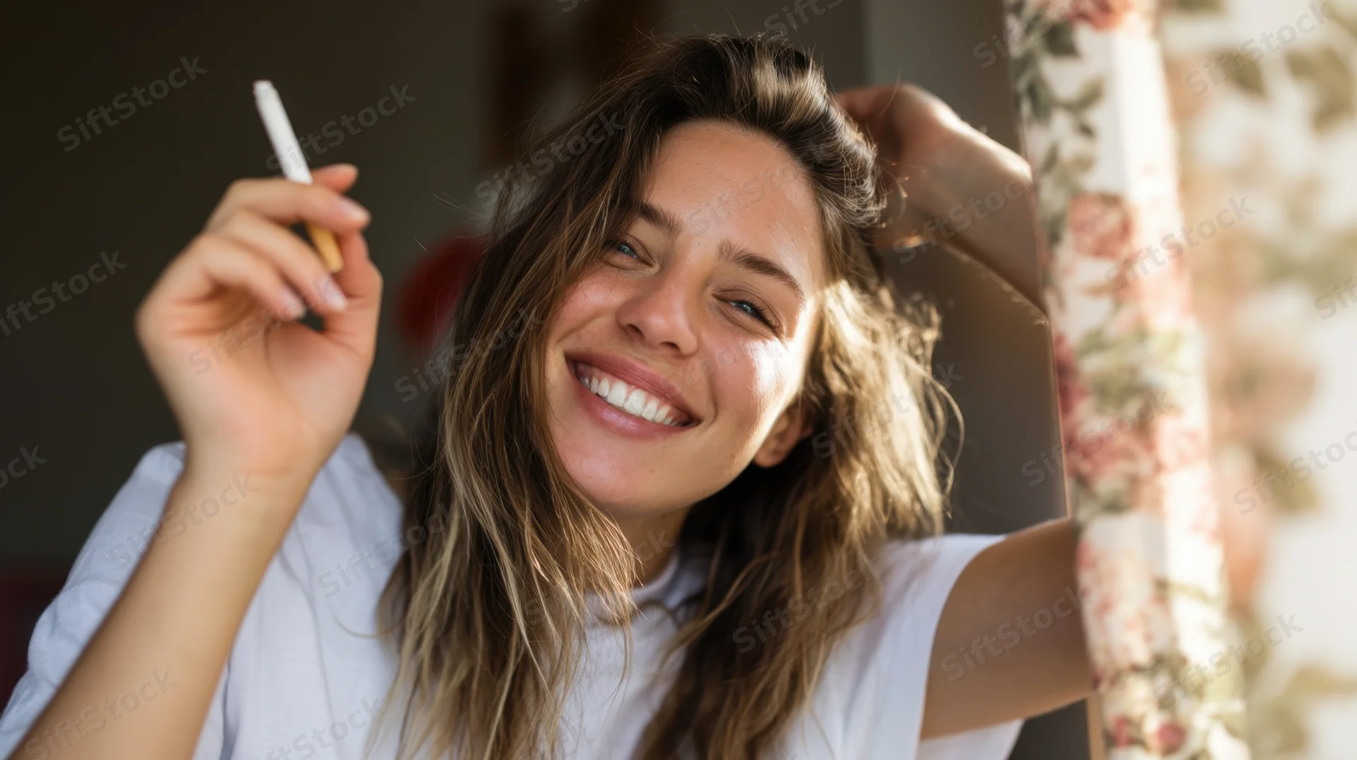 Woman smiling while holding a cigarette indoors near a floral curtain