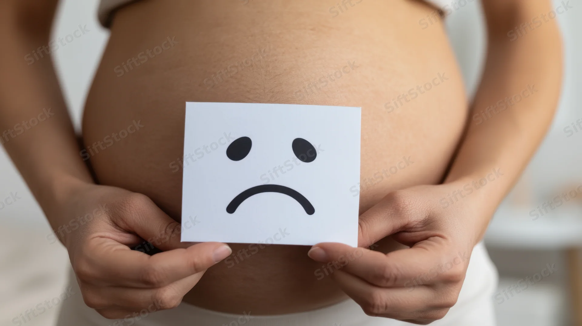 Pregnant woman holding sad face card over her abdomen indoors