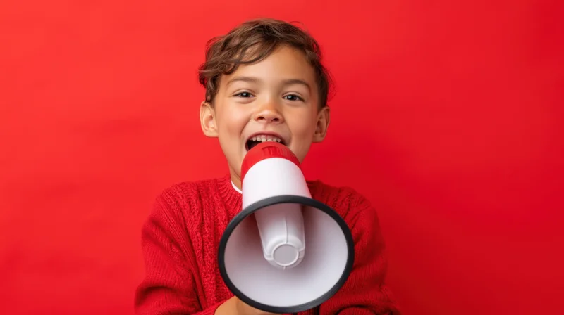 Young hispanic boy shouting into a red and white megaphone against a red background