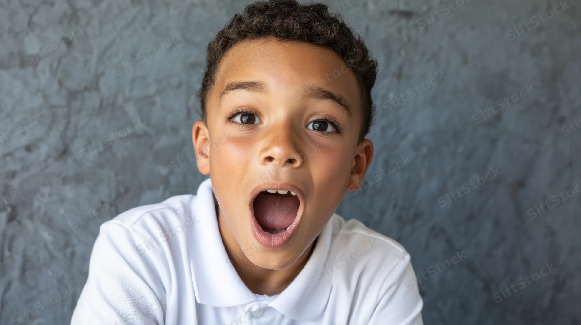 Young boy with wide eyes and open mouth showing surprise against a gray textured wall