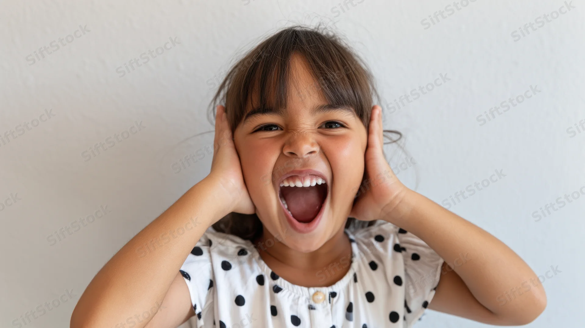 Young hispanic girl laughing with hands on ears wearing white polka dot dress indoors