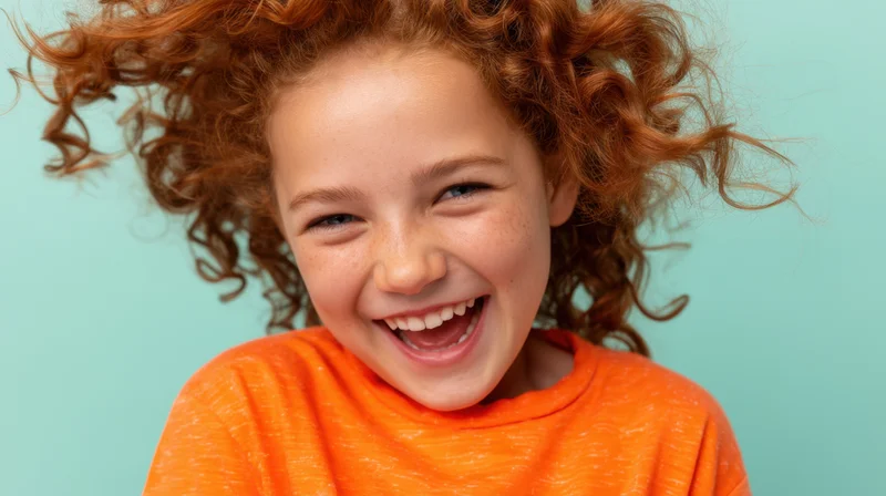 Young girl with vibrant curly red hair laughing joyfully in orange shirt against teal background
