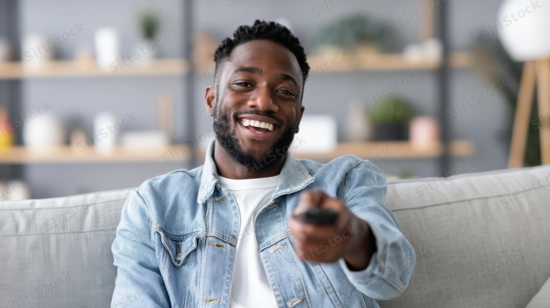 Young black man smiling while holding remote on light gray couch in living room